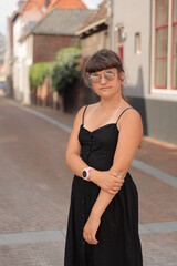Fototapeta premium A young beautiful girl wearing in round glasses and black dress standing in city background in Holland, outdoor shoot
