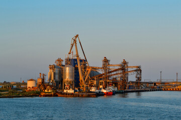 Coastal industrial seascape with red cranes and metal structures with port and transport ship. Generic view of industrial harbor with vessels docked in sunset light