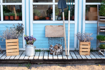 Facade blue home with garden tools: shovel, pitchfork, rake, firewood and pots flowers. Cozy Autumn decor veranda house. Gardening. Wooden porch of house with plants, logs, dry flowers. Exterior patio