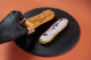 Raspberry eclair and caramel eclair held by hand in black glove on round black rock stand. Eclairs...