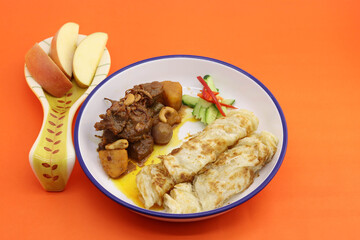 food served in round plate on orange background. in the plate have massaman beef curry and roti and sliced apple served at side of food.