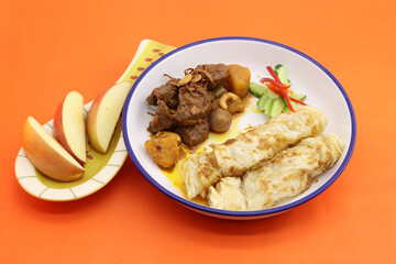 food served in round plate on orange background. in the plate have massaman beef curry and roti and sliced apple served at side of food.