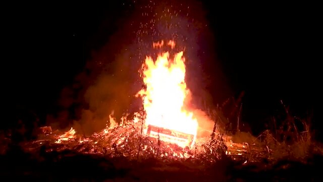 Huge solstice campfire burning in the black of night. Wideshot. What exactly is in there? A cupboard?