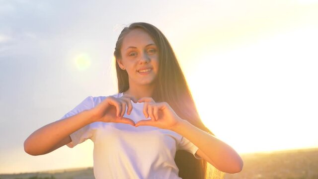 A girl at sunset looks into the camera and makes a heart symbol out of her fingers. Heart shape with hands, a sign of love. Concept of health care, vacation, help, Valentine's day