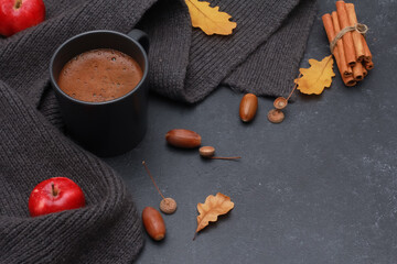Black cup of coffee, oak leaves, acorns, red apple, grey scarf on dark background. Copy space, high angle. Autumn background, fall season, thanksgiving, coffee shop menu concept