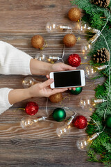 Female hands with a smartphone among the New Year's decorations and a Christmas tree on a wooden background. Choosing gifts online for the new year
