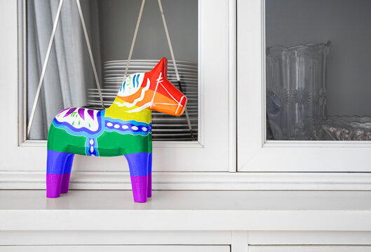 Dalecarlian horse (Dalahäst) in LGBTQ rainbow color version. Hand painted wooden statue of a horse in front of white sideboard or buffet with dishes. Concept of acceptance and inclusivness.