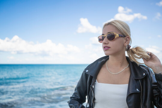 Outdoor, day. Portrait of a young Italian woman with long blond hair tied in a ponytail with a white low-cut top enriched by a delightful white mother-of-pearl necklace, sunglasses and black jacket