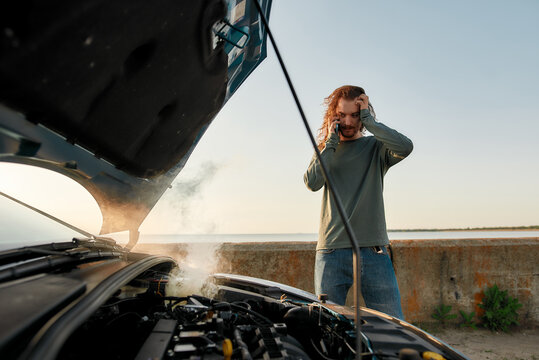 Young man looking desperate, using smartphone, calling for assistance while standing near his broken car with open hood