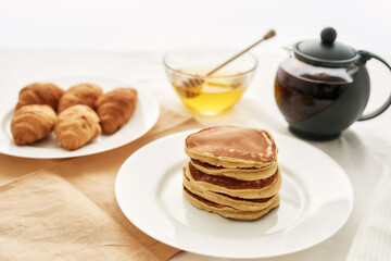 Close up of a stack of sweet tasty pancakes, croissants, honey in a bowl with dipper and a teapot, Breakfast served on the table over white background