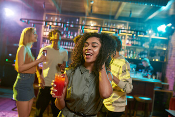 Attractive mixed race young woman smiling at camera while posing with a cocktail in her hand and friends chatting, having drinks at the bar counter in the background