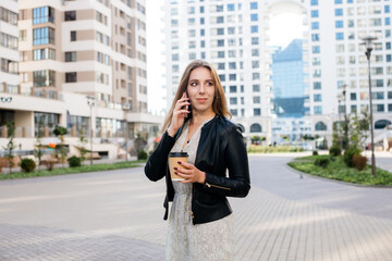 Fototapeta premium Beautiful woman is speaking on the phone with a cup of coffee at the city street background.