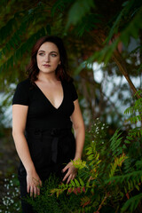 Stunning young woman poses for photo in late summer in elevated urban park - standing in greenery in black blouse and pants