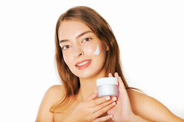 Close up image of young beautiful woman applying facial cream to face, white background, holding jar with moisturizer