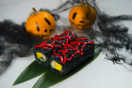Halloween Holiday Dish Food Art concept. Japanese Black Rice Sushi Roll with red spider web decoration on top. Two Scary painted pumpkins with black spiders on background. Roll served on bamboo leaves