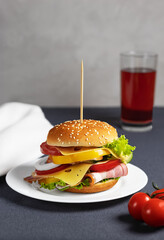 Homemade burger with double cheese, bacon slices, tomatoes, bell peppers, onion rings and lettuce on a white plate close-up.