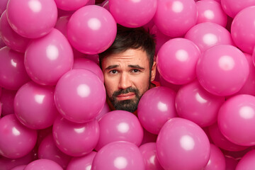 Fototapeta na wymiar Photo of dissatisfied bearded European man being tired after arrangement birthday party poses around many pink air balloons makes photo over creative background. Holiday celebration concept.