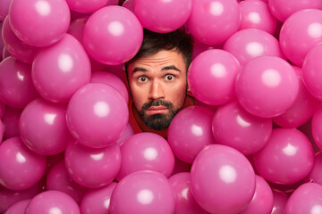 Impressed bearded European guy poses over decorated photo zone sticks out head through inflated pink balloons has fun on birthday party makes funny shot stares bugged eyes at camera. Celebration time