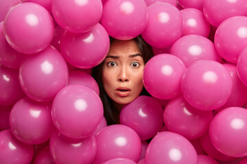 Fototapeta na wymiar Shocked puzzled young Asian lady looks through many pink inflated balloons surprised by arranged party hears shocking news. Brunette woman spends free time on birthday party or festive event