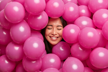 Fototapeta na wymiar Positive cheerful young woman giggles positively with closed eyes and smiles broadly surrounded by rosy air balloons celebrates birthday spends free time on graduation party has childish mood.