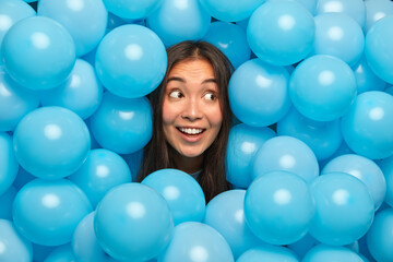 Happy ethnic woman looks mysteriously aside and broad smile poses against blue balloons awaits for special event. Cheerful Asian girl sticks out head through decorated background celebrates birthday