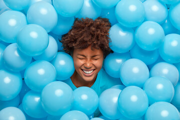 Fototapeta na wymiar Overjoyed curly haired Afro American woman feels amused and entertained on party has fun and sticks out head through wall decorated with blue balloons expresses happy emotions. Festive event