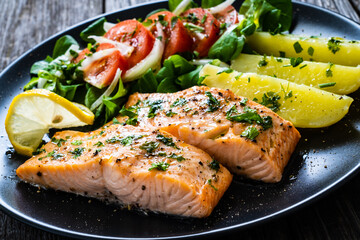 Fried salmon fillet with boiled potatoes, lemon and vegetable salad served on black plate on wooden table
