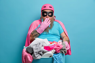 Indoor shot of stupefied bearded superhero man holds laundry basket covers mouth wears rubber gloves cape and mask stunned to have household duties isolated on blue background. Housework concept