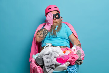 Plump bearded husband in superhero costume helps wife with work about house holds basin full of...