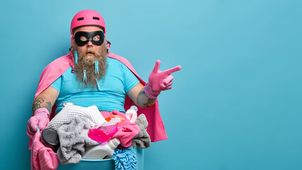 Studio shot of shocked bearded man in hero costume points on blank space for your advertisement poses against basin full of laundry isolated on blue background. Cleaning service and household - obrazy, fototapety, plakaty