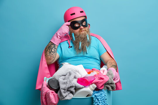 Thoughtful bearded man poses with basin full of laundry thinks how to wash everything rubs temple and looks pensively aside dressed in super hero costume. Housework and domestic chores concept