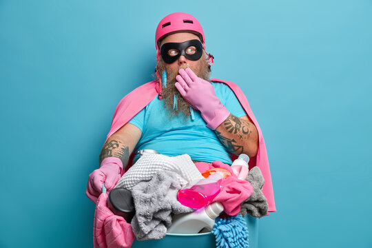 Photo of scared frightened man dressed in super hero costume poses against basin full of laundry wears rubber protective gloves poses against blue background. Everyday routine and housework.