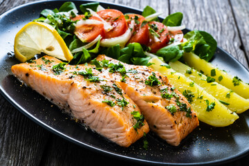Fried salmon fillet with boiled potatoes, lemon and vegetable salad served on black plate on wooden table
