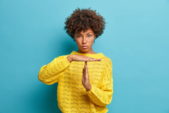 Serious self confident curly haired Afro American woman makes timeout gesture demonstrates limit asks to stop dressed in yellow knitted jumper isolated on blue background. Body language concept