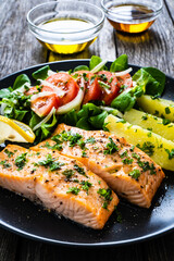 Fried salmon fillet with boiled potatoes, lemon and vegetable salad served on black plate on wooden table
