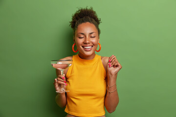 Happy pleased dark skinned woman closes eyes and smiles toothily raises clenched fist and drinks cold cocktail has happy mood dressed in casual outfit poses in studio against green background