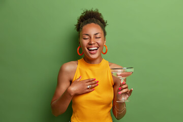 Overjoyed dark skinned woman laughs happily keeps eyes closed and smiles broadly has fun at party drinks cocktail expresses positive emotions wears yellow shirt poses over vivid green background - Powered by Adobe