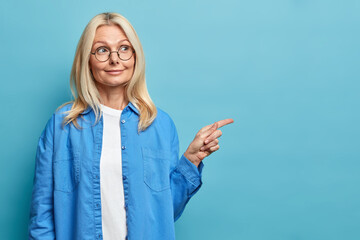 Cute wrinkled woman with blonde hair points on copy space aside shows something interesting has...