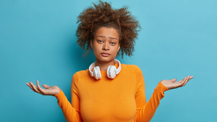 Indifferent teenage girl has bushy curly shrugs shoulders and looks with careless facial expression...