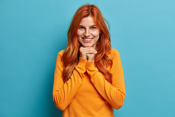 Portrait of pleasant looking redhead young woman smiles happily and keeps hands under chin looks directly at camera with satisfied expression dressed in casual wear isolated on blue background