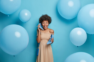 Excited fashionable dark skinned woman with curly hair holds high heel shoes pretends talking via phone waits for important event dressed in beige evening dress poses over decorated background