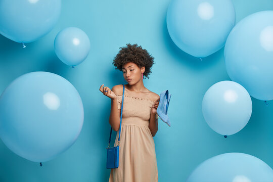 Serious fashionable woman looks at her new manicure dressed in beige dress carries blue high heel shoes to fit bag dresses for special occasion surrounded with inflated balloons over blue background
