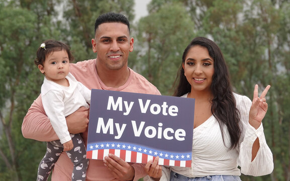 A Young Hispanic Family Smiles For The Camera While Holding Up A Sign Saying 