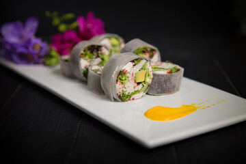 Macro of No Rice Sushi Roll with crab meat (sticks), salad leaves, cream cheese, tobiko caviar wrapped in daikon radish on white Japanese plate served with yellow painting brush stroke and flowers

