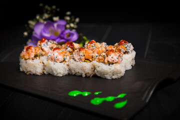 Side view of Japanese Sushi Roll with salmon on top served with sauce on black Asian serving plate. Green painting brush strokes and violet flowers on dark background. Creative serving dish art
