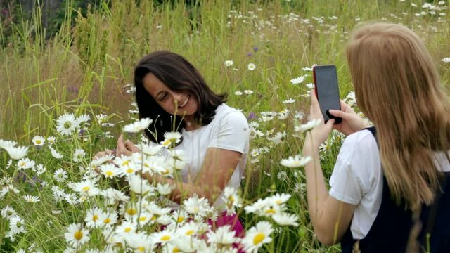 Two happy young girls on a chamomile field.Friends take photos on their phones, laugh and inhale the scent of wild meadow flowers while enjoying a summer day. Chamomile field. Holidays