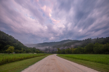 Dirt road through mountains and forest on a cloudy foggy day between grass fields 