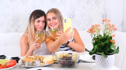  friendship and party concept - two young beautiful girlfriends drink wine and take selfie on smartphone in a cozy kitchen at the table, selective focus