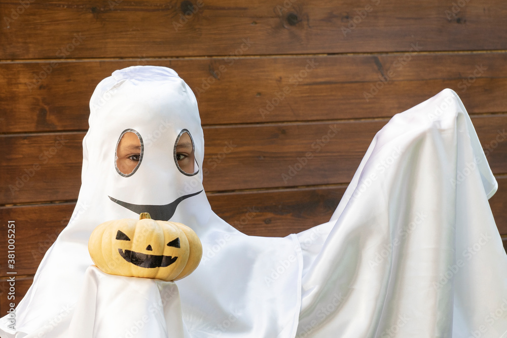 Wall mural unrecognizable boy in white costume of halloween ghost holds decorated pumpkin, on brown wooden back