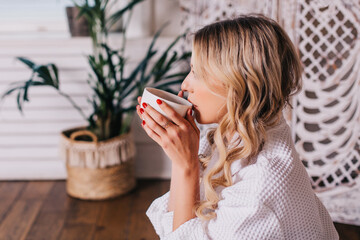 a young woman sits on a bed in the morning at home in a white bathrobe and drinks coffee. quarantined girl woke up late in the morning